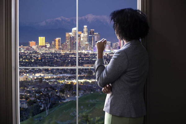 Black female business woman looking out the window of an office in Los Angeles.  She looks like a female architect thinking of urban development or a city mayor or governor planning zoning laws. 