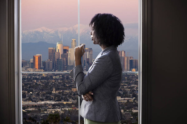 Black female business woman looking out the window of an office in Los Angeles.  She looks like a female architect thinking of urban development or a city mayor or governor planning zoning laws. 