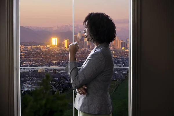 Black female business woman looking out the window of an office in Los Angeles.  She looks like a female architect thinking of urban development or a city mayor or governor planning zoning laws.