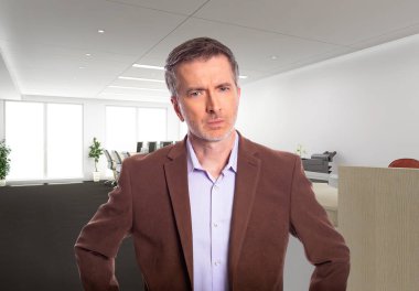 Middle-Aged businessman with gray hair and wearing a brown jacket standing in an office.  Depicts startup and corporate business.  He is looking upset or angry.