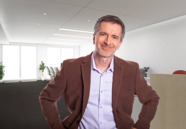 Middle-Aged businessman with gray hair and wearing a brown jacket standing in an office.  Depicts startup and corporate business.  He is smiling and happy.