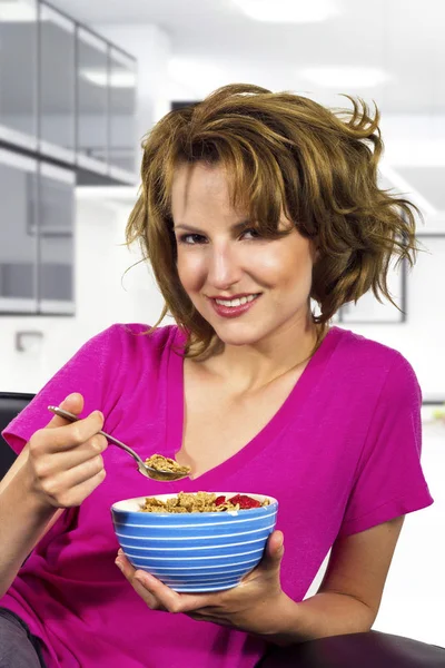 Caucasian female with frizzy hair sitting on a couch eating a breakfast bowl of cereal.  She is at home and looks like she just woke up and doing a morning routine.