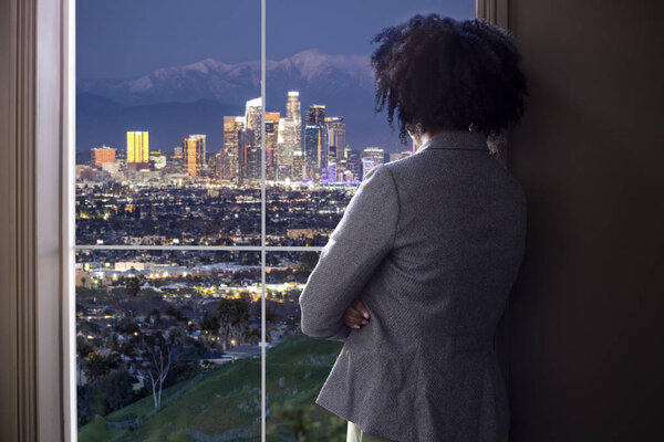 Black female businesswoman looking worried or tired by an office window with a view of downtown Los Angeles, California.  She looks like a start-up business owner or a politician or city planner architect. 