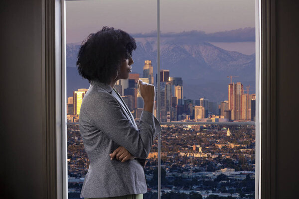 Black female businesswoman looking worried or tired by an office window with a view of downtown Los Angeles, California.  She looks like a start-up business owner or a politician or city planner architect. 