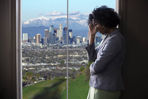 Black female businesswoman looking worried or tired by an office window with a view of downtown Los Angeles, California.  She looks like a start-up business owner or a politician or city planner architect. 