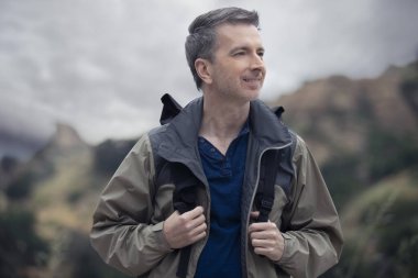 Orta yaşlı beyaz erkek yürüyüşçü dağlarda tek başına yürüyor. Bulutlu gökyüzü huzurlu ve adam mutlu görünüyor. Açık hava aktivitelerini ve seyahat turizmini gösteriyor.