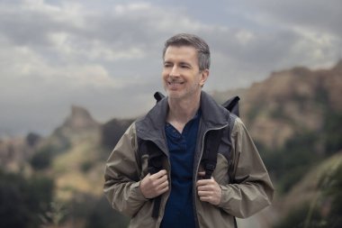Orta yaşlı beyaz erkek yürüyüşçü dağlarda tek başına yürüyor. Bulutlu gökyüzü huzurlu ve adam mutlu görünüyor. Açık hava aktivitelerini ve seyahat turizmini gösteriyor.