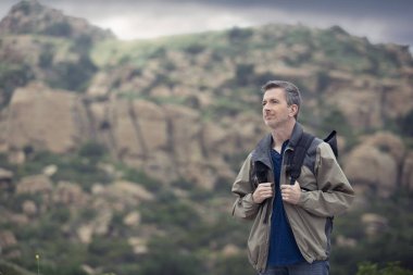 Orta yaşlı beyaz erkek yürüyüşçü dağlarda tek başına yürüyor. Bulutlu gökyüzü huzurlu ve adam mutlu görünüyor. Açık hava aktivitelerini ve seyahat turizmini gösteriyor.