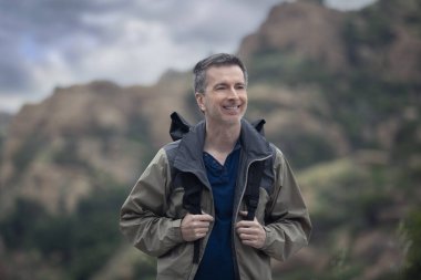 Orta yaşlı beyaz erkek yürüyüşçü dağlarda tek başına yürüyor. Bulutlu gökyüzü huzurlu ve adam mutlu görünüyor. Açık hava aktivitelerini ve seyahat turizmini gösteriyor.