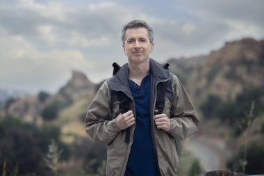 Orta yaşlı beyaz erkek yürüyüşçü dağlarda tek başına yürüyor. Bulutlu gökyüzü huzurlu ve adam mutlu görünüyor. Açık hava aktivitelerini ve seyahat turizmini gösteriyor.