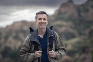 Orta yaşlı beyaz erkek yürüyüşçü dağlarda tek başına yürüyor. Bulutlu gökyüzü huzurlu ve adam mutlu görünüyor. Açık hava aktivitelerini ve seyahat turizmini gösteriyor.