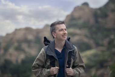 Orta yaşlı beyaz erkek yürüyüşçü dağlarda tek başına yürüyor. Bulutlu gökyüzü huzurlu ve adam mutlu görünüyor. Açık hava aktivitelerini ve seyahat turizmini gösteriyor.