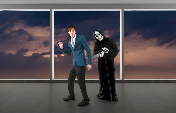 Man in a Halloween grim reaper ghost costume chasing, mocking and making fun of scared businessman running away.  Can also depict death following a man as a metaphor for life insurance.