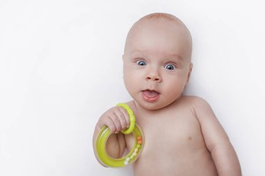 Yeni doğan bebek elinde oyuncakla beyaz bir arka planda yatıyor ve şaşkınlıkla kameraya bakıyor. Ağzıyla kameraya şaşkınlıkla bakıyor. 