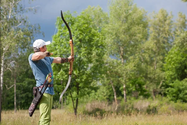 A man in a blue T-shirt and cap shoots an archery. He trains in the woods. — Stock Photo, Image