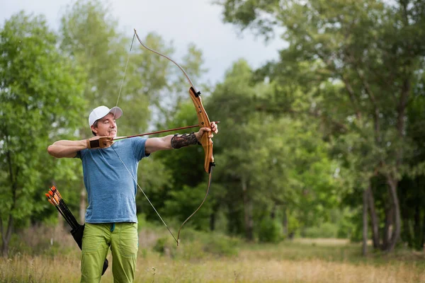 A man in a blue T-shirt and cap shoots an archery. He trains in the woods. — Stock Photo, Image