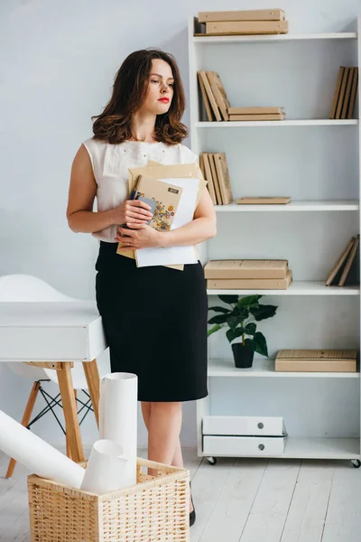 Una chica con una blusa blanca está de pie con su teléfono y libros en sus manos en la oficina. Ella tiene el pelo castaño recortado debajo del quad . —  Fotos de Stock