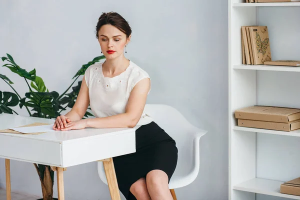 Ένα κομψό κορίτσι με λευκό πουκάμισο και μαύρη φούστα κάθεται σε ένα λευκό τραπέζι. Σκέφτηκε. Το κορίτσι κάθεται σε ένα γραφείο διακοσμημένο σε λευκό και μπεζ χρώμα. — Φωτογραφία Αρχείου