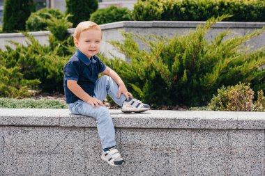 cute boy 2-3 years old sits on a stone parapet near a green bush.