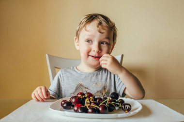 A three-year-old child sits in the kitchen of the house and eats cherries. he had a sly look aside. 