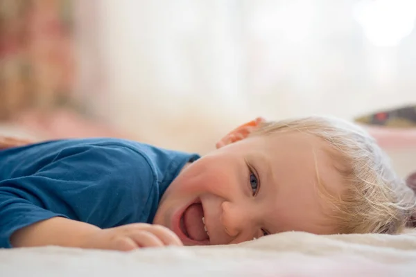 The little one in the blue jacket is lying on the bed and laughing. Little gay boy lies on the bed — Stockfoto