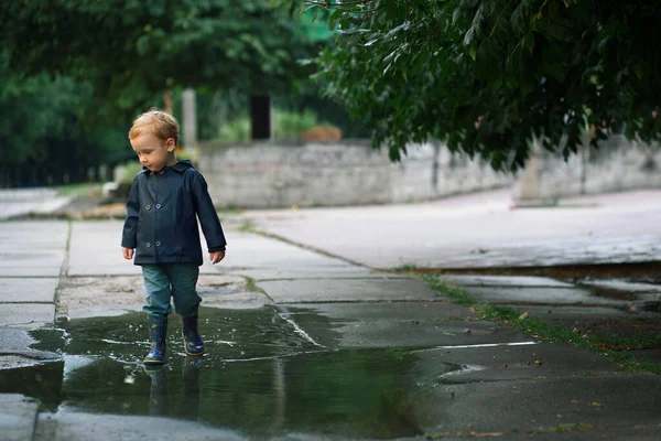 Un garçon en imperméable et bottes en caoutchouc marche sur une flaque d'eau . — Photo