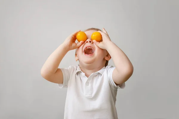 Un chaval muy divertido se deleita con la fruta naranja, se pone la fruta en los ojos y se ríe . — Foto de Stock