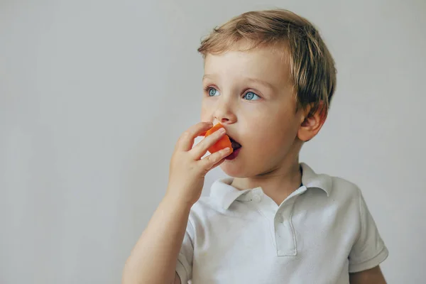 Um menino todler em uma camiseta branca tem uma fruta laranja . — Fotografia de Stock