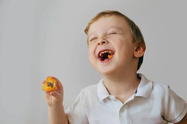 Um menino de três anos ri depois de morder uma deliciosa fruta de laranja. O rapaz está a usar uma t-shirt branca — Fotografia de Stock