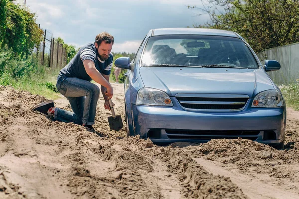 A blue car stuck in the sand.. A strong man with a beard digs a shovel of the wheel of the car.