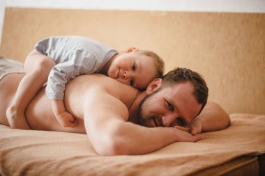 Küçük çocuk babasının üzerine yatıyor. Çocuk ve baba evdeki yatakta yatıyorlar.. 