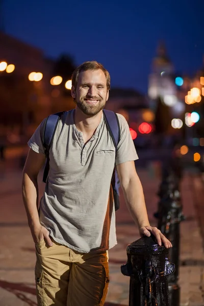 Молодой человек с бородой стоит на ночной площади и смеется. Парень гуляет по летнему ночному городу . — стоковое фото