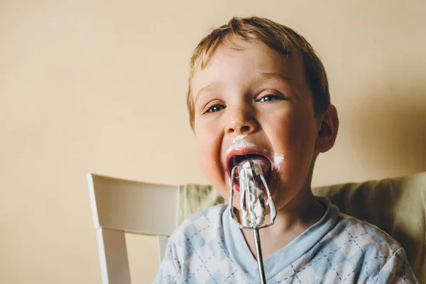 Um pequeno menino pré-escolar senta-se em uma cadeira contra uma parede bege e lambe sua língua com um creme branco de um batedor. Criança manchada com creme — Fotografia de Stock