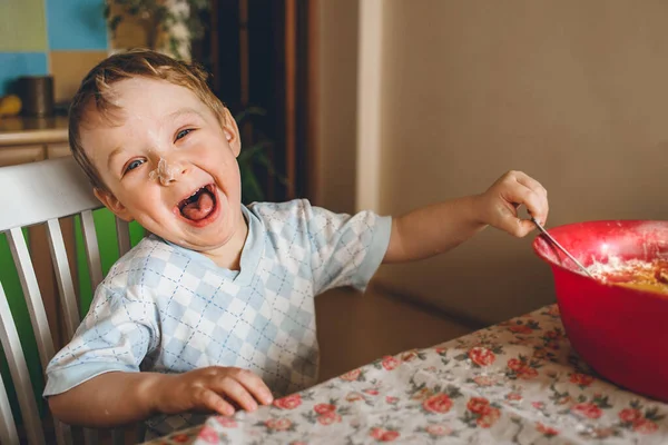 O menino se senta na cozinha à mesa e ri da câmera. A criança ajuda sua mãe a cozinhar uma torta. Mexe a massa numa tigela. — Fotografia de Stock