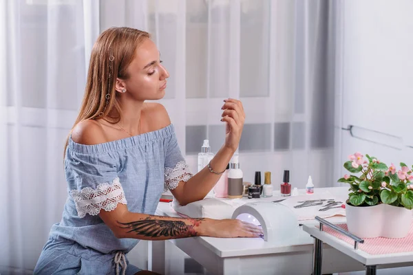 Vacker flicka med tatuering sitter vid bordet och torkar under LED-lampan färsk manikyr och nagellack — Stockfoto