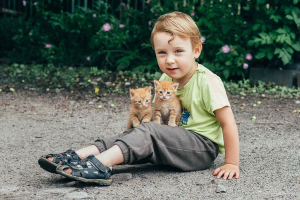 Niño pequeño bebé se sienta en la calle y sostiene dos gatitos rojos —  Fotos de Stock