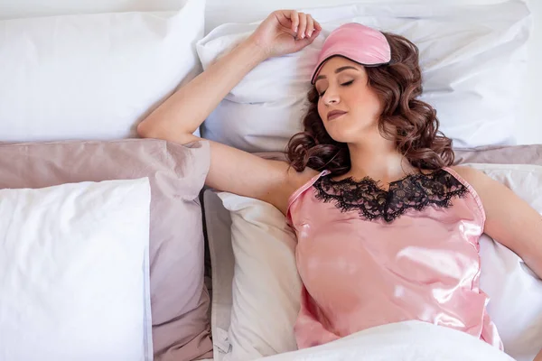 Una mujer joven en ropa de dormir rosa y una máscara de dormir se encuentra en la cama sobre almohadas dormidas —  Fotos de Stock