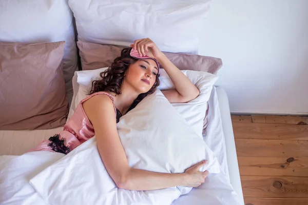 Una joven en ropa de dormir rosa y una máscara de dormir se encuentra en la cama abrazando almohadas —  Fotos de Stock