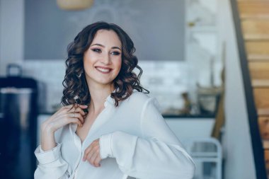 Makyajlı ve saç stili kıvırcık saçlı genç bir kadının portresi. Mutfakta oturmuş gülümsüyor ve yan tarafa bakıyor.