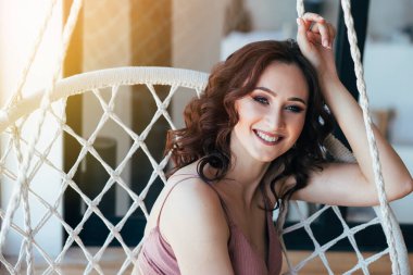 Makyajlı ve bukleli genç bir kadın hasır bir sandalyede oturur ve gülümser. 