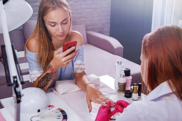 Caucasian blonde girl with a phone in her hand sits in the salon and makes a manicure at the master. — Stock Photo, Image