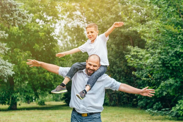 Een man met een baard rolt op de schouders van een tienerjongen in een park. Pa en zoon spelen vliegtuigen in de natuur., — Stockfoto