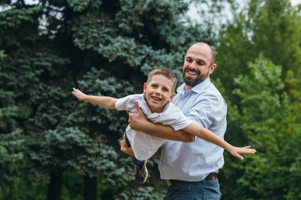 Un uomo barbuto rotola sulle spalle di un adolescente in un parco. Papà e figlio giocano aerei in natura, — Foto Stock