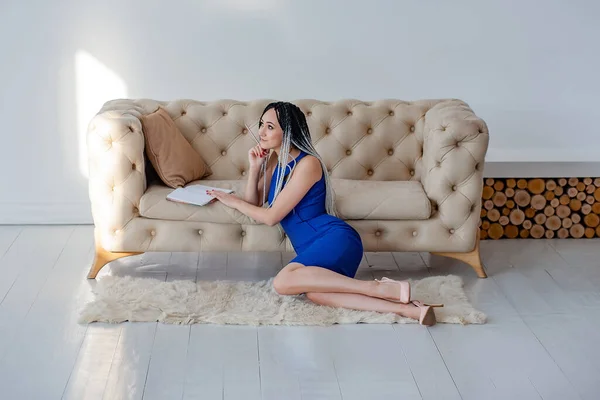 Piękna dziewczyna w sukience siedzi na podłodze w pobliżu kanapy i pisze notatki w notatniku — Zdjęcie stockowe