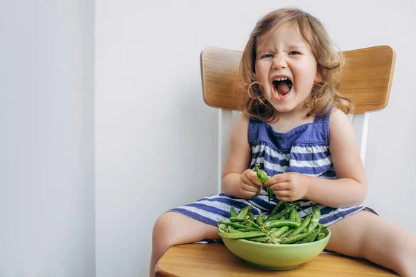 Alegre pouco todler menina em um vestido listrado comer ervilhas e sorrir — Fotografia de Stock