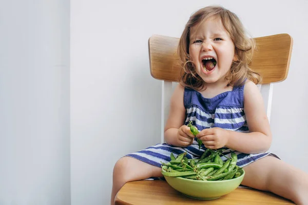 Allegro piccolo bambino ragazza in un abito a righe mangiare piselli e smorfie Fotografia Stock