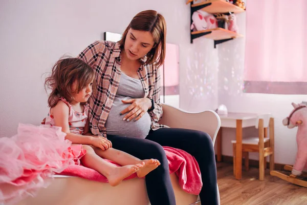 Kaukasiska gravid kvinna sitter på sängen med en småbarn flicka och pratar. Stockbild