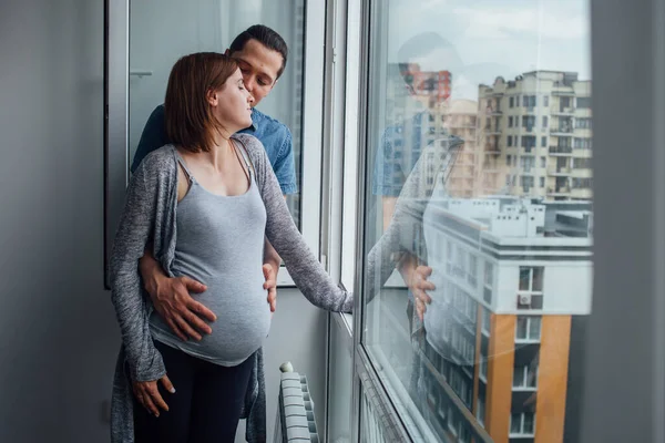 En man och en gravid kvinna står hemma nära ett öppet fönster och pratar. Maken kramar sin fru, de stannar hemma och tittar på gatan från fönstret. Stockfoto