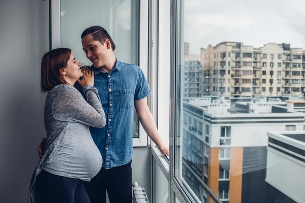 En man och en gravid kvinna står hemma nära ett öppet fönster och pratar. Maken kramar sin fru, de stannar hemma och tittar på gatan från fönstret. Stockbild