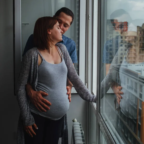 En man och en gravid kvinna står hemma nära ett öppet fönster och pratar. Maken kramar sin fru, de stannar hemma och tittar på gatan från fönstret. Royaltyfria Stockfoton